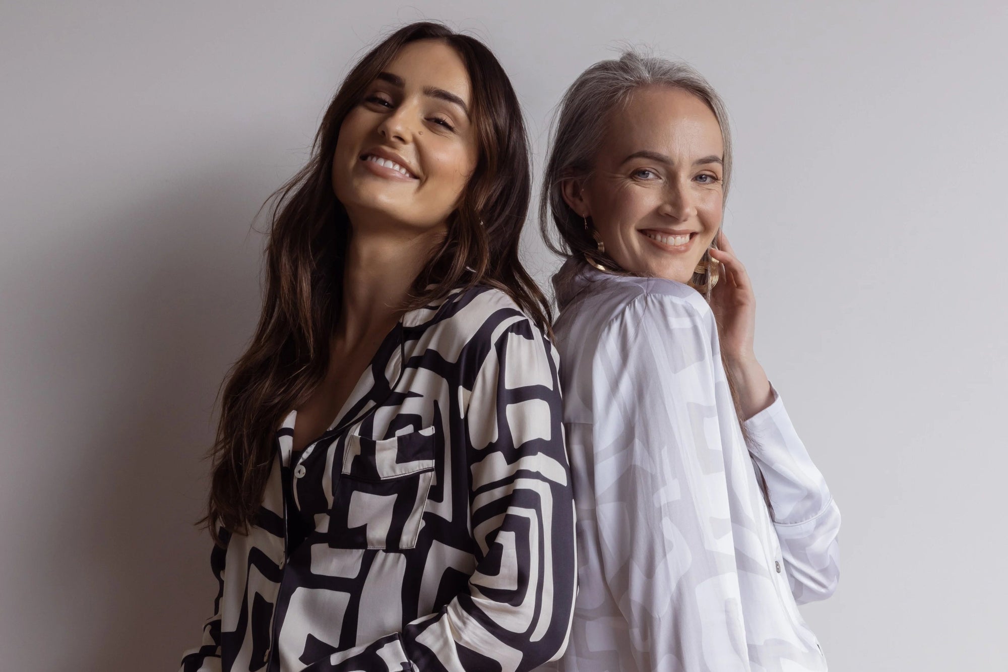 Two women standing back to back smiling, wearing beautiful bamboo pj sets with a bold geometric pattern, one in black and cream, the other in white and grey colours.