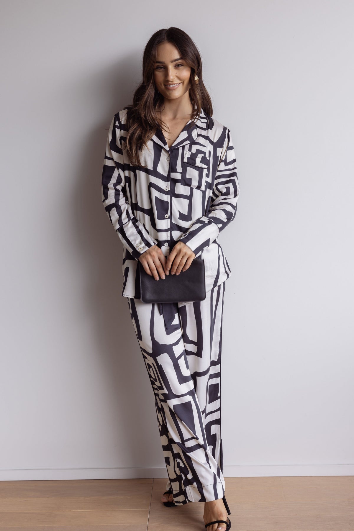 Woman wearing silky, breathable 100% bamboo PJ set showing a long sleeve pyjama top with button up design, pants with pockets and bold geometric print in black and cream. Dressed up with a bag and heels.