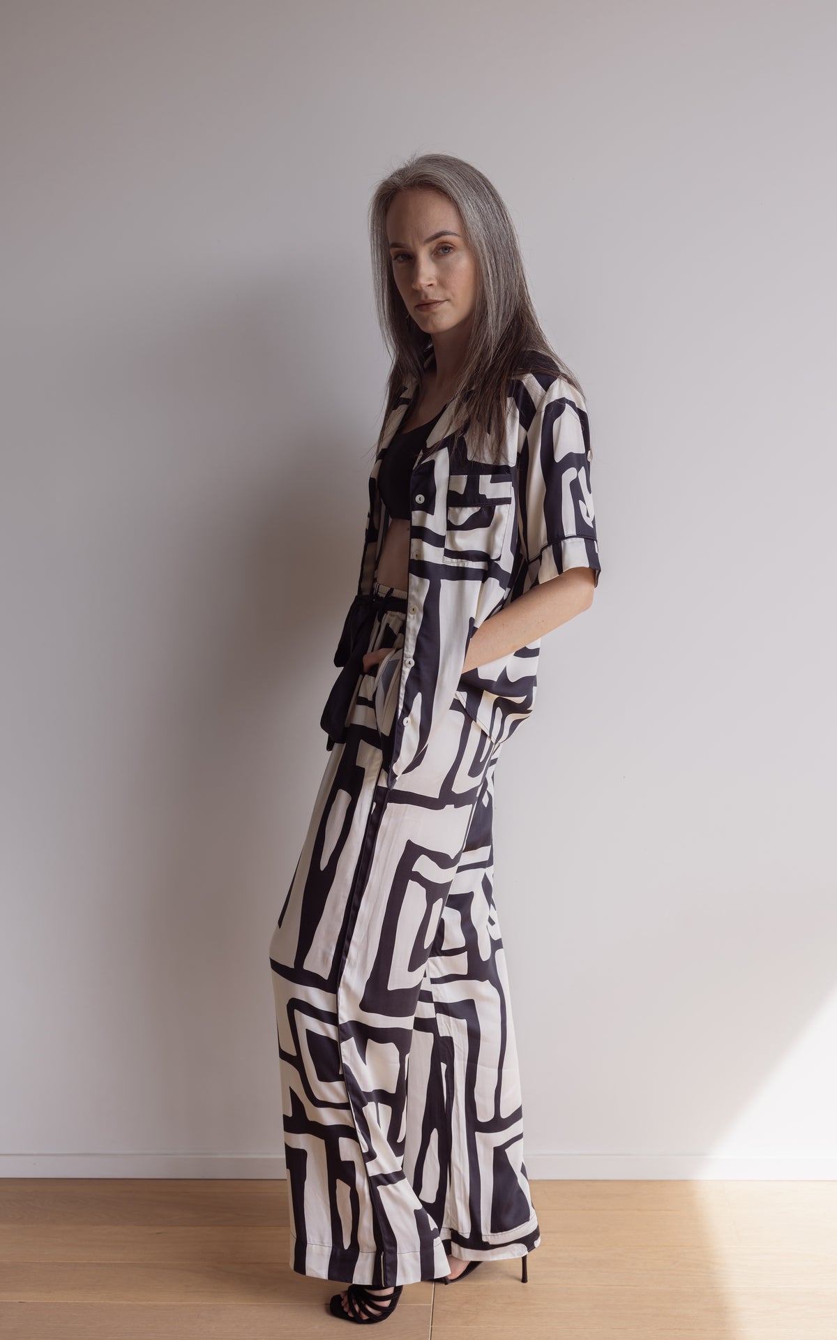 Woman standing  to the side in short sleeve pyjama top and pants made from silky bamboo. In a bold geometric print, black and cream colour.