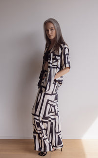 Woman standing  to the side in short sleeve pyjama top and pants made from silky bamboo. In a bold geometric print, black and cream colour.