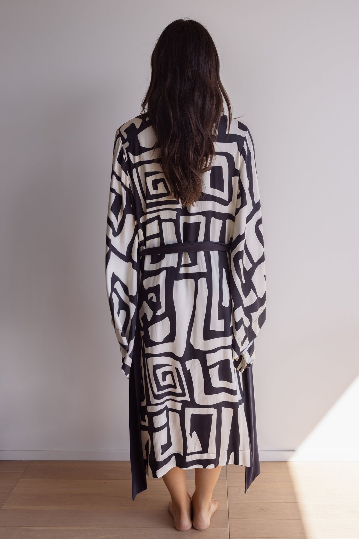 Woman wearing a kimono-style robe. Made from silky-soft bamboo with a flowing fit, featuring a unique print design and a tie waist for an adjustable fit. Hidden pockets. Perfect for lounging. Black and cream colour.