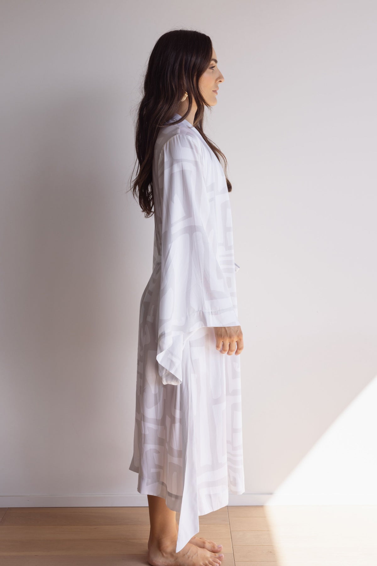 Woman wearing a comfortable kimono-style robe. Made from silky-soft, breathable bamboo. Featuring a unique print design in light grey and white, with and a tie waist for an adjustable fit and hidden pockets. Perfect for relaxing.