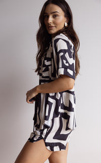 Woman wearing a comfortable and breathable bamboo short pyjama set. Including a short-sleeve button-up top and matching shorts with a relaxed fit, pockets and elasticated waistband. Geometric print in black and cream.