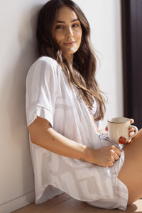 Woman sitting, wearing a comfortable and breathable bamboo short pyjama set. Including a short-sleeve button-up top and matching shorts, pockets and elasticated waistband. Geometric print in white and grey.